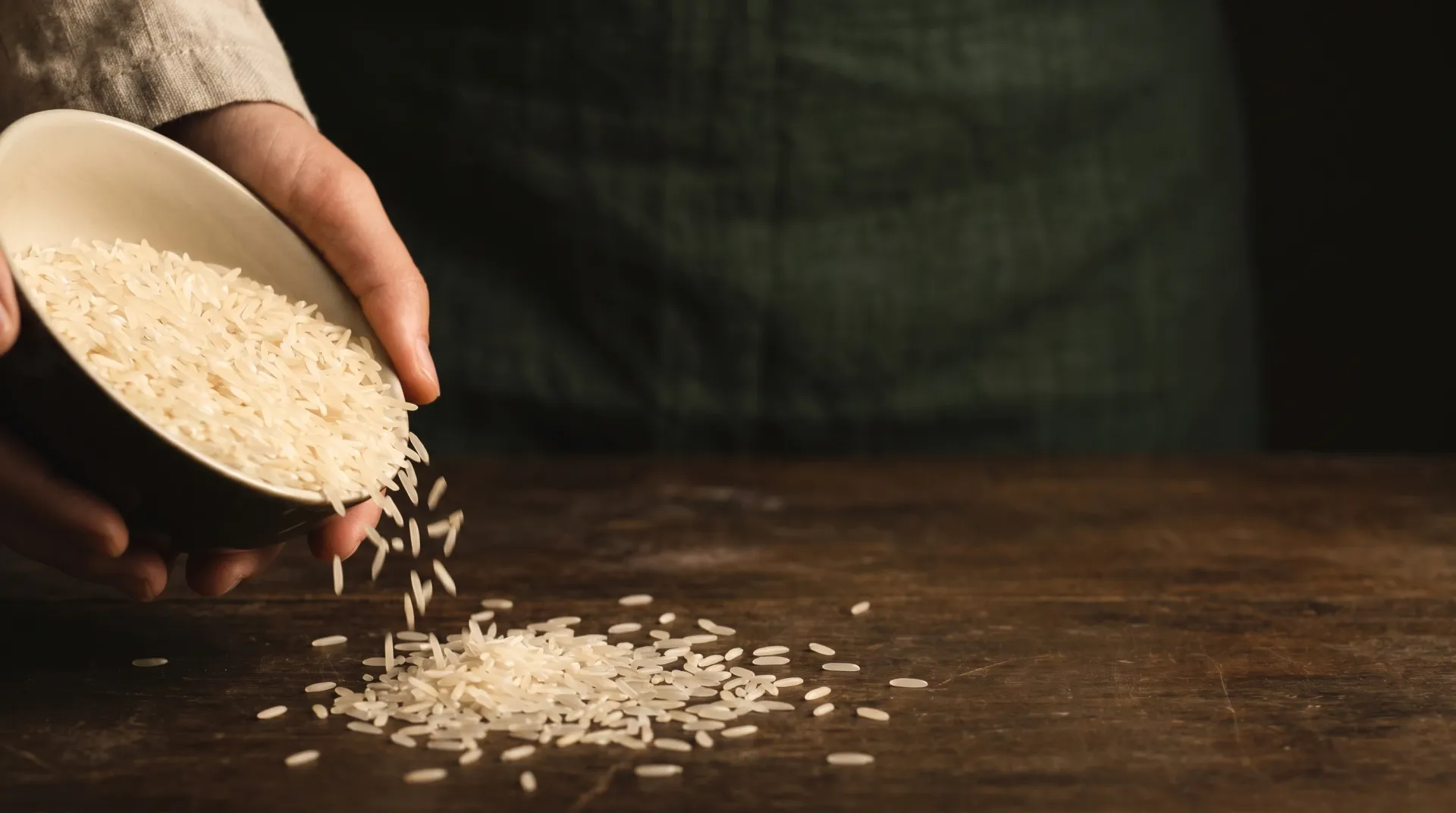 Premium long-grain basmati rice poured from a cream bowl onto aged dark wood, against a deep paddy-green ground